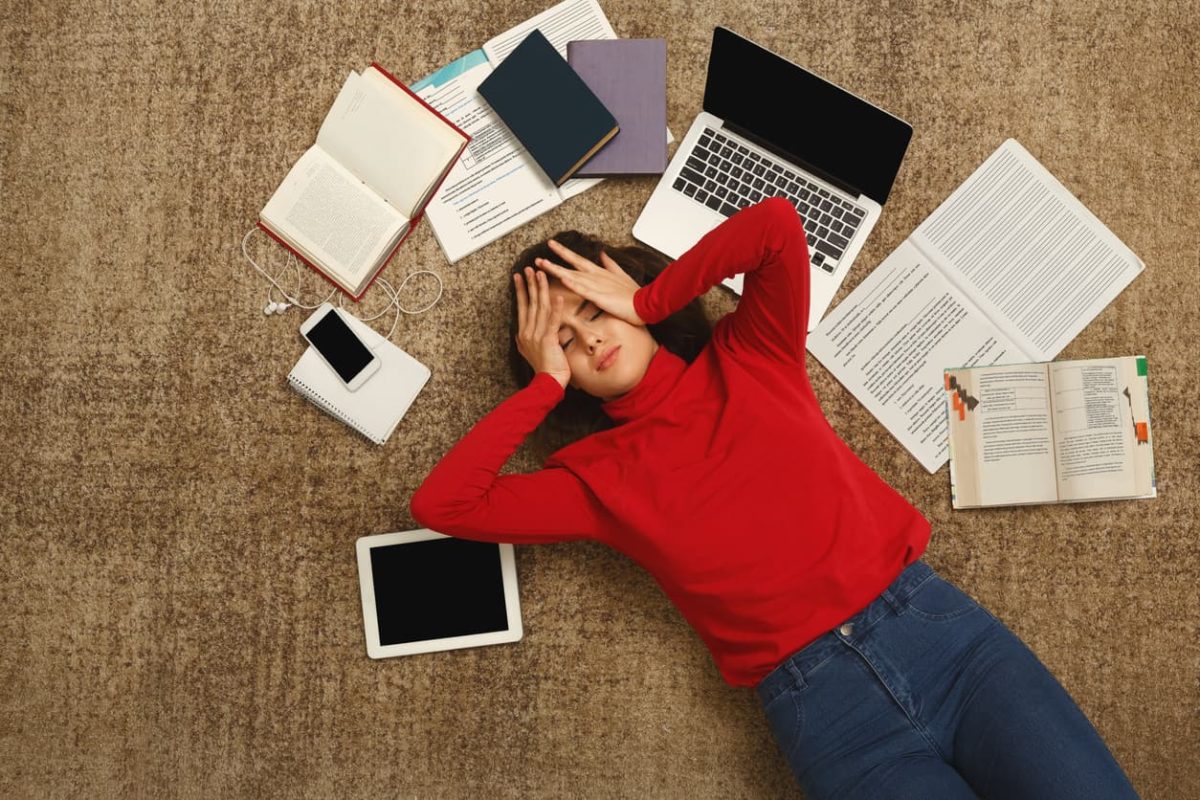 Exhausted student girl lying on the floor among textbooks, tests and gadgets, copy space. Woman holding head with hands, got tired while preparing for exams. Education and overworking concept